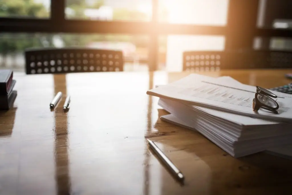This is a photo of a desk with a stack of papers. There are three pens on the desk and a pair of glasses resting on top of the papers.